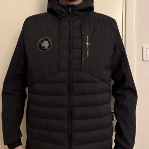 Svart pufferjacka från Sail Racing - Svart pufferjacka från Sail Racing med huva och snygga logodetaljer på bröstet. Jackan har quiltad framsida, dragkedja hela vägen och långärmade ärmar. Perfekt för kalla dagar med sin stilrena look och sportiga känsla.