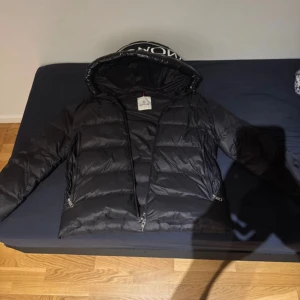 Svart Moncler dunjacka med huva - Säljer en svart Moncler dunjacka med stor huva och tydlig logga på kragen. Jackan har två dragkedjefickor, quiltad design och klassisk Moncler-märke på ärmen. Perfekt för kalla dagar och har en riktigt snygg streetstil.