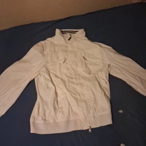 Vit vindjacka från ZNY - Säljer en fräsch vit vindjacka från De Blasio med hög krage och dragkedja framtill. Jackan har två bröstfickor med dragkedja, ribbad nederkant och ett snyggt De Blasio-märke på ärmen. Perfekt för dig som gillar clean och sportig stil.