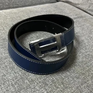 Marinblått skärp med H-spänne - Snyggt marinblått bälte med silverfärgat H-format spänne. Bältet har vita kontrastsömmar längs kanterna och är gjort i slätt skinn. Perfekt accessoar för att lyfta din outfit med en stilren detalj.