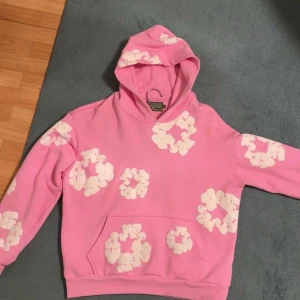 Rosa hoodie från Denim Tears med blommor - Säljer en rosa hoodie från Denim Tears med vita blommiga tryck över hela tröjan. Tröjan har huva och magficka, perfekt för dig som vill sticka ut med en cool och färgglad stil. Skön passform och riktigt snygg design!