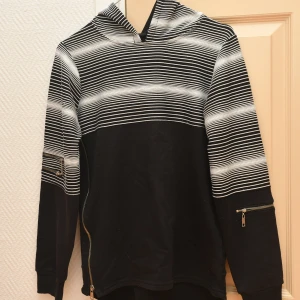 Svartvit randig hoodie med dragkedjor - Cool svart hoodie med vita ränder upptill och på ärmarna. Tröjan har huva och snygga dragkedjor på ärmarna och sidan. Perfekt för dig som gillar streetstyle och vill sticka ut lite extra. Mjuk insida och långärmad modell.