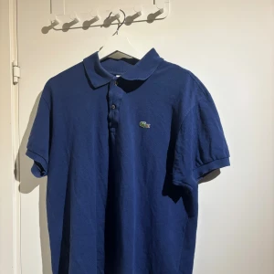 Blå pikétröja från Lacoste - Snygg blå pikétröja från Lacoste med klassisk krage och två knappar framtill. Tröjan har korta ärmar och den ikoniska gröna krokodilloggan på bröstet. Tillverkad i mjuk bomull som känns skön mot huden. Perfekt för dig som gillar stilren och sportig look.