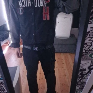 Svart bomberjacka från Hoodrich - Säljer en svart bomberjacka från Hoodrich med stora röda och vita bokstäver på bröstet. Jackan har knappar framtill och ribbade muddar vid ärmslut och nederkant. Perfekt för dig som gillar streetwear och vill sticka ut med coola detaljer.