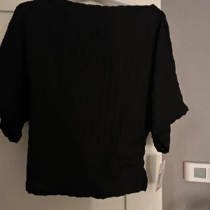 Svart strukturerad topp från Nelly - Svart topp från Nelly i storlek medium med snygg strukturerad yta. Modellen har korta ärmar och en loose passform, perfekt för dig som gillar en enkel men stilren look. Materialet känns mjukt och har en lätt textur som ger extra stöd dimension till outfiten.