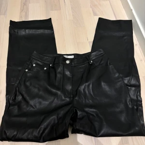 Svarta raka byxor i skinnimitation - Snygga svarta byxor från NA-KD i skinnimitation med klassisk femficksdesign och raka ben. Byxorna har hög midja, silverfärgade nitar och knapp, samt bälteshällor.