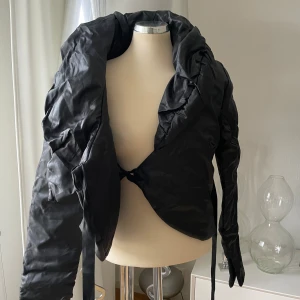 Svart omlott pufferjacka - Svart pufferjacka från MF Italy med stor krage och omlottstängning med knytband i midjan. Jackan är croppad och har långa ärmar med breda muddar. Materialet är 80% polyester och 20% nylon, vilket ger en glansig och lätt känsla. Perfekt för dig som gillar statement-plagg.Oanvänd men har lite ljusare streck vid kragen 