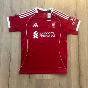 Liverpool röd fotbollströja Adidas M - Liverpool FC matchtröja i röd färg från Adidas, med vita detaljer och tryck. Kortärmad modell med rund hals, broderat klubbmärke och sponsorloggor. Materialet är lätt och andas, perfekt för träning eller match. Rygg med namn 'ISAK' och nummer 9.