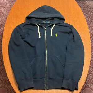Marinblå hoodie från Polo Ralph Lauren - Snygg marinblå hoodie från Polo Ralph Lauren med dragkedja, vita snören och klassisk gul logga på bröstet. Tröjan har huva, kängurufickor och är tillverkad i mjuk bomull. Perfekt för chill dagar eller när du vill ha en clean streetstyle.
