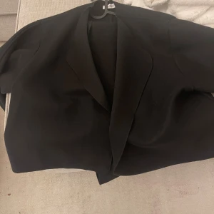 Svart omlottkavaj med bälte - Säljer en svart kavaj från Wikipe med omlottmodell och tillhörande bälte i midjan. Kavajen har en stilren design med breda slag och rak passform. Perfekt för dig som gillar minimalistisk och clean stil.