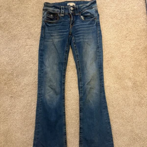 Blå bootcut jeans från Gina Tricot - Snygga blå jeans från Gina Tricot i storlek 152 med klassisk bootcut passform. Jeansen har fem fickor, orangea sömmar och en medelblå tvätt. Perfekta för dig som gillar retrovibbar och vill ha ett par jeans med lite utsvängda ben.
