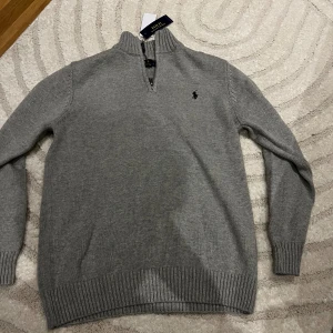 Grå stickad tröja från Polo Ralph Lauren - Snygg grå stickad tröja från Polo Ralph Lauren med halv dragkedja och ribbade muddar. Klassisk broderad logga på bröstet och hög krage. Perfekt för lager-på-lager och passar till många olika stilar.