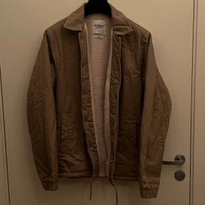 Beige coachjacka från Dobber - Snygg beige coachjacka från Dobber med mjukt teddyfoder på insidan. Jackan har klassisk krage, snörning nedtill och tryckknappar framtill. Perfekt för dig som gillar stilren och enkel design med lite extra värme. Jackan är i bra skick då den har bara vart ihop knycklad i garderoben de senaste 3 åren.