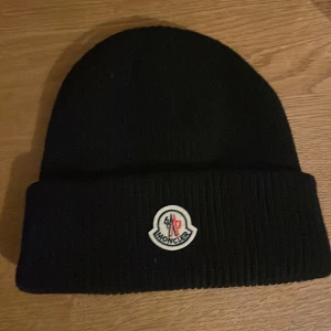 Svart Moncler mössa i ribbstickad ull - Svart ribbstickad mössa från Moncler med klassisk uppvikt kant och broderad Moncler-logga framtill. Snygg och enkel design som passar perfekt till kalla dagar. Tillverkad i mjuk ull för extra värme och komfort.