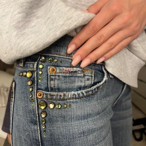 Blå bootcut jeans med stenar - Snygga blå bootcut jeans med coola gröna och gula stenar längs fickorna. Jeansen har låg midja, klassiska bakfickor med broderad detalj och är tillverkade i stretchig denim. Perfekta för dig som vill sticka ut med lite extra bling. Jag är 167 och säljer de för att de är liiite för korta! Ungefär st 34, skriv för en bild med måtten. 