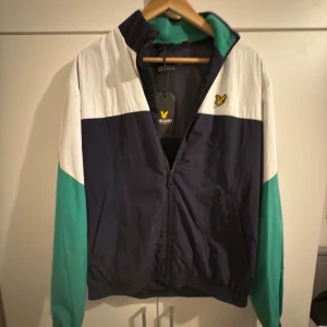 Lyle & Scott färgblockad vindjacka - Priset kan diskuteras. Snygg vindjacka från Lyle & Scott med färgblock i vitt, mörkblått och turkos. Jackan har dragkedja framtill, ståkrage och det klassiska gula fågel-loggan broderad på bröstet. Perfekt för dig som gillar sportig och retro stil.