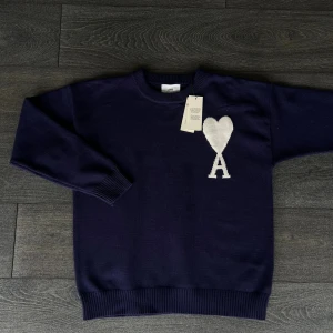 Mörkblå Ami Paris tröja - Snygg mörkblå stickad tröja från Ami med ett vitt hjärta och bokstaven A broderat på bröstet. Tröjan har rund hals, ribbade muddar och långärmad passform. Perfekt för dig som gillar stilrena detaljer och vill ha något som sticker ut.