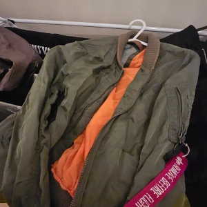 Grön bomberjacka med orange foder - Säljer en klassisk grön bomberjacka med orange foder och ribbad krage. Jackan har dragkedja framtill och en ficka med dragkedja på ärmen. Perfekt för dig som vill ha en cool och tidlös streetstil.