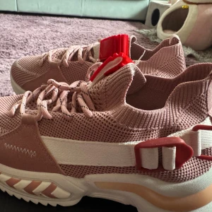 Rosa chunky sneakers strl 39, helt nya. - Säljer ett par snygga chunky sneakers i ljusrosa med vita och röda detaljer. Skorna har stickad ovandel, snörning och coola plastdetaljer på sidan. Sulan är tjock och har mönstrade inslag i rosa och vitt. Perfekt för dig som gillar streetstyle och vill sticka ut.