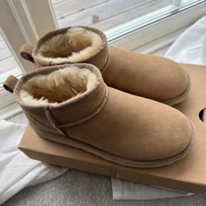 UGG Ultra Mini boots i sand - Säljer ett par UGG Ultra Mini boots i färgen sand med mjukt fårskinnsfoder och mocka på utsidan. De har rund tå, platt sula och UGG-logga bak. Perfekta för kalla dagar och riktigt sköna att glida runt i. Snygg och enkel design som passar till allt. Impregnerade och använda ungefär 3 gången, inga defekter. nypris 2099