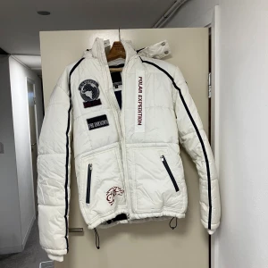 Vit pufferjacka från Smog med patchar - Säljer en vit pufferjacka från Smog med coola patchar och broderier, svart piping längs ärmarna och huva med broderad detalj. Jackan har två dragkedjefickor, snörning nedtill och texten 'Polar Expedition' framtill. Perfekt för kalla dagar.