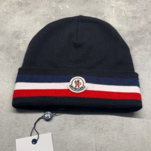 Moncler mössa med ränder - Svart Moncler mössa i mjuk ull med breda blå, vita och röda ränder runt kanten. Klassisk Moncler-logga framtill. Perfekt för dig som vill ha en stilren och sportig look med premiumkänsla.