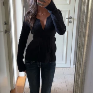Svart omlott kofta med knytband - Säljer en svart stickad omlott kofta med knytband i midjan och djup v-ringning. Koftan har lång ärm och sitter snyggt figurnära. Perfekt att slänga över ett linne eller topp för en chill men ändå stilren look.