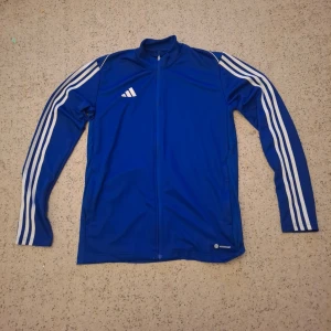 Blå Adidas långärmad ziptröja - Säljer en blå långärmad Adidas ziptröja med vita ränder längs ärmarna och Aeroready-tryck. Tröjan har hel dragkedja framtill, klassisk Adidas-logga på bröstet och är tillverkad i ett lätt, sportigt polyestermaterial. Aldrig använd.