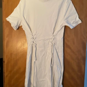 Vit kortklänning med snörning - Säljer en vit kortklänning med snörningsdetaljer i midjan. Klänningen har korta ärmar och är tillverkad i mjukt bomullsmaterial. Enkel och stilren design som ger en cool och avslappnad vibe.