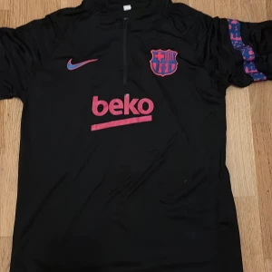 Svart FC Barcelona fotbollströja Nike - Säljer en svart FC Barcelona fotbollströja från Nike med rosa och blå detaljer. Tröjan har korta ärmar med ett coolt mönster i blått och rosa på ärmarna, halv dragkedja framtill och broderat klubbmärke. Materialet är lätt och andas, perfekt för träning. Ingår även byxor 
