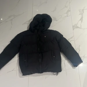 Svart pufferjacka från GANT med huva - Svart pufferjacka från GANT med stor, skön huva och dragkedja framtill. Jackan har två fickor med knappstängning och GANT-logga på bröstet. Perfekt för kalla dagar och har en clean, modern look som funkar till streetwear. Hade ett hål men sydde ihop de märks inte ens eller syns 