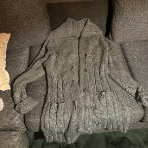Grå stickad kofta med knäppning - Mysig grå stickad kofta med grovt kabelstickat mönster och stora fickor framtill. Koftan har lång ärm, hög krage och snygga träknappar som stängning. Storlek står inte men passar för mig som har M och tror typ den skulle passa L o S med bara sitta lite olika.