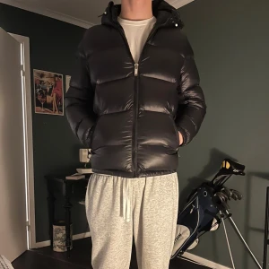 Svart pufferjacka från Zara - Svart pufferjacka från Zara med huva och två sidofickor med dragkedja. Jackan har en quiltad design och hel dragkedja framtill. Materialet är glansigt syntet och jackan är långärmad med en loose passform. Perfekt för kalla dagar när du vill hålla värmen nu inför vintern! Köpte den för 1300 och säljer för halva priset! Yttermaterial: 100 % nylon, fyllning: 100% plyester, foder: 100% polyester, vadderingstyp: varm vaddering