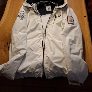 Vit vindjacka Team Adventure M - Snygg vit vindjacka från Team Adventure med coola broderade patchar och tryck på ärmen. Jackan har flera dragkedjefickor, elastisk nederkant och huva. Perfekt för dig som gillar sportig stil och detaljer som sticker ut.