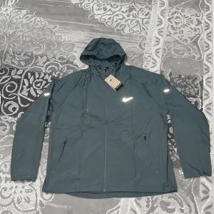 Mörkgrön Nike vindjacka med huva - Snygg mörkgrön vindjacka från Nike med huva och reflekterande detaljer. Jackan har dragkedja framtill, två fickor med dragkedja och meshpaneler på insidan. Perfekt för dig som gillar sportig stil och vill ha något lätt och funktionellt.