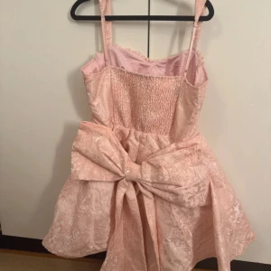 Rosa kort klänning med stor rosett - Supersöt ljusrosa kort klänning med breda axelband och en stor rosett framtill. Klänningen har ett blommigt mönster i tyget och är tillverkad i glansig satin. Smockad rygg och spetsdetaljer vid bysten ger extra charm. Perfekt för dig som vill sticka ut med en romantisk look.