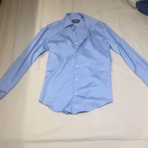 Ljusblå slim fit skjorta Dressmann - Snygg ljusblå skjorta från Dressmann i slim fit-modell. Skjortan är tillverkad i bomullsstretch för extra komfort och har klassisk krage samt vita knappar framtill. Perfekt för dig som gillar en stilren och fräsch look.