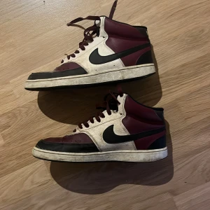 Nike skor - Nike Air Force  sneakers i vinrött, svart och vitt med klassisk svart swoosh på sidan. Skorna har snörning, högt skaft och platt sula med cirkulärt mönster undertill. Materialet är skinn och syntet, och färgtonen är mörk vinröd med kontrasterande detaljer. De är i bra skick och inte använda mycket. Storlek 42 box finns om man vill ha den också! Vid snabb affär kan jag gå ner i pris!
