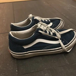 Blå Vans Old Skool sneakers - Säljer ett par klassiska Vans Old Skool sneakers i mörkblått med vita detaljer och snörning. Skorna har en vit sula, rund tå och den ikoniska sidoremsan. Ovandelen är i mocka och textil, med Vans-logga på hälen. Perfekt för dig som gillar streetstyle.