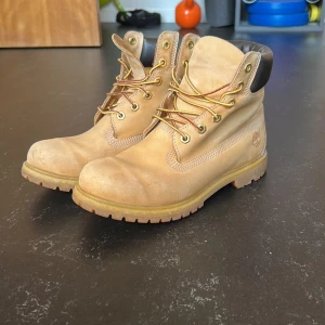 Beiga Timberland boots  - Klassiska Timberland boots i beige nubuckskinn med robust snörning och grov sula. Skorna har vadderad svart krage runt ankeln och ikoniska Timberland-loggan på sidan. Perfekta för höst och vinter. Använda men i väldigt bra skick. Nypris 1499. 
