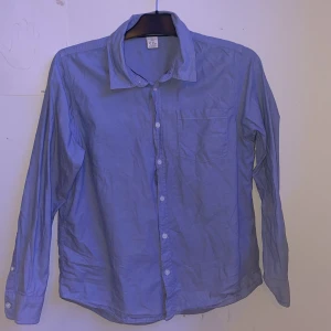 Blå skjorta från Lindex, stl 164 - Klassisk blå skjorta från Lindex med lång ärm, bröstficka och knappar framtill. Skjortan är i bomull och har en stilren, enkel design som passar perfekt till jeans eller chinos.