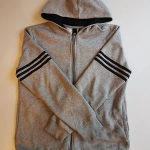 Grå Adidas hoodie med dragkedja - Grå hoodie från Adidas med svart logga och text på ryggen samt klassiska svarta ränder på ärmarna. Tröjan har huva, hel dragkedja framtill och ribbade muddar. Perfekt för chill eller träning.