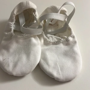 Vita balettskor från Bloch - Säljer ett par klassiska vita balettskor från Bloch med rund tå och elastiska band över foten. Skorna är tillverkade i bomull och har en mjuk, flexibel sula som passar perfekt för dans. Enkla att ta på och av tack vare resår vid öppningen.
