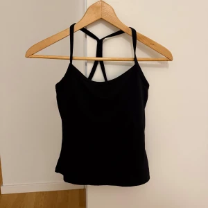 Svart linnetopp med snygga axelband - Svart linnetopp med smala axelband som korsas i ryggen för en sportig och cool look. Toppen är ärmlös och har en enkel, stilren design som passar perfekt till gymmet eller andra träningspass. Materialet känns mjukt och följsamt mot kroppen.