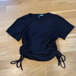 Svart ribbad topp med snörning från H&M - Säljer en svart ribbad topp från H&M Divided i storlek S. Toppen har korta ärmar och cool snörning på båda sidorna som ger en snygg, justerbar passform. Materialet är mjukt och stretchigt, perfekt för att skapa en chill och trendig look.
