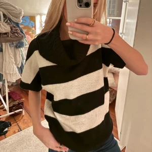 Randig polotröja i svart och vitt - Säljer en snygg polotröja med breda svarta och vita ränder. Tröjan har korta ärmar och en stor, mysig krage. Stickad i mjukt material som känns skönt mot huden. Perfekt för dig som gillar en chill och trendig stil.