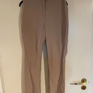 Beige kostymbyxor från NA-KD, stl 34 - Stilrena beige kostymbyxor från NA-KD Reborn i rak modell. Byxorna har diskreta bakfickor och är tillverkade i ett mjukt polyestertyg. Perfekta för en clean och snygg look.