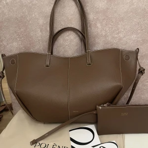 Brun axelväska från Polène - Stilren brun axelväska från Polène i slätt skinn med vita kontrastsömmar. Väskan har både handtag och axelrem samt medföljande matchande clutch. Perfekt för dig som vill ha en rymlig och snygg väska till vardagen.