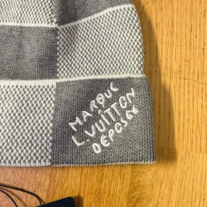 Gråvit rutig mössa Loui buitton - Snygg grå och vit rutig mössa från Loui buitton med broderad text 'Marque L. Vuitton Deposee' framtill. Mössan har klassisk passform och är tillverkad i mjukt stickat material. Perfekt accessoar för att lyfta din vinterstil.