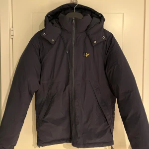 Mörkblå pufferjacka från Lyle & Scott - Snygg mörkblå pufferjacka från Lyle & Scott med huva och gul broderad logga på bröstet. Jackan har dragkedja framtill, snörning vid huvan och praktiska fickor. Perfekt för kalla dagar och har en clean, modern look.